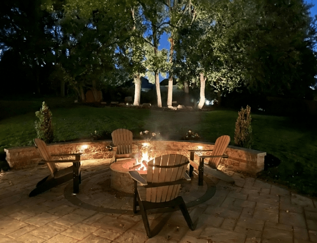 A paver patio with custom fire pit and sitting wall accented by outdoor lighting and landscaping.